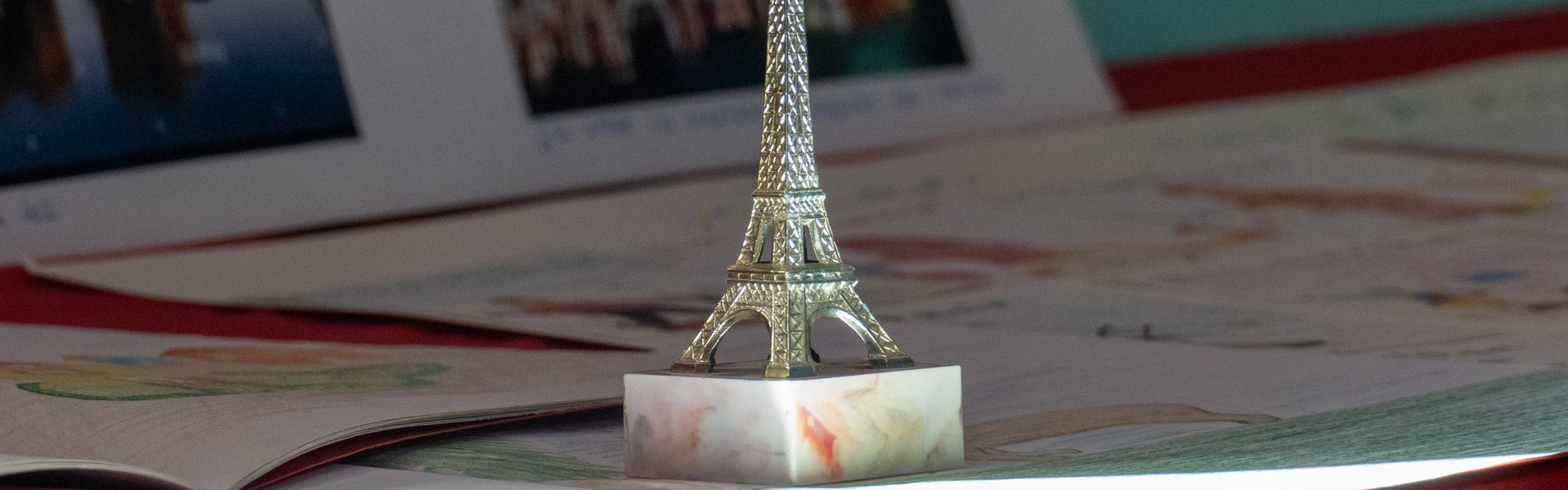 Eine kleine Eiffelturm-Figur steht auf einem Marmorsockel, umgeben von Papieren und Fotos auf einer roten Fläche, während Sonnenlicht die Szene in den Glanz taucht.