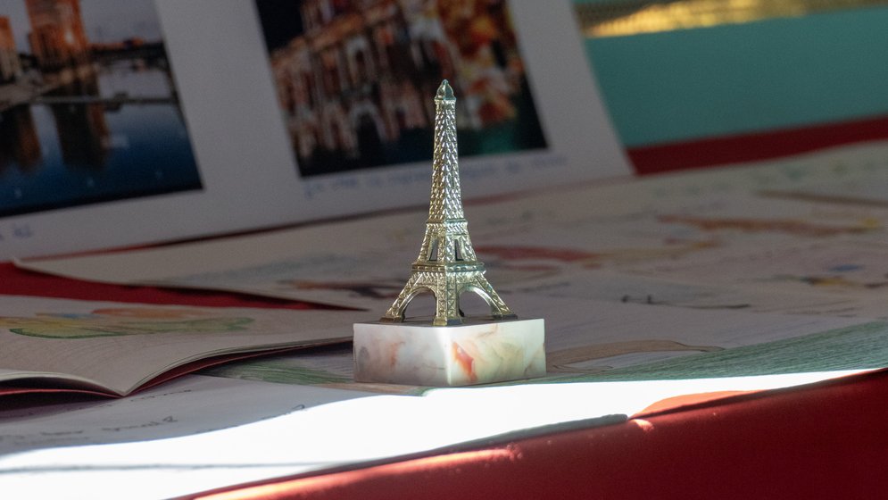 Eine kleine Eiffelturm-Figur steht auf einem Marmorsockel, umgeben von Papieren und Fotos auf einer roten Fläche, während Sonnenlicht die Szene in den Glanz taucht.