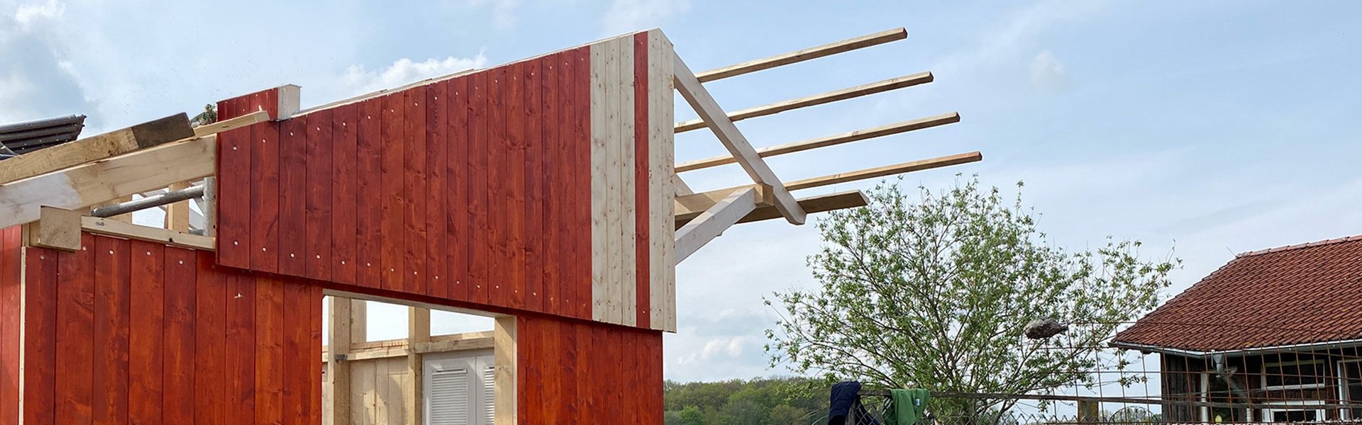 Ein noch nicht ganz fertiggestellter Holzschuppen mit roter Verkleidung, freiliegenden Balken und überall verstreutem Werkzeug. Frisch gestrichene Bretter trocknen auf Sägeböcken. Im Hintergrund sind ein Maschendrahtzaun und ein Haus mit rotem Dach zu sehen.