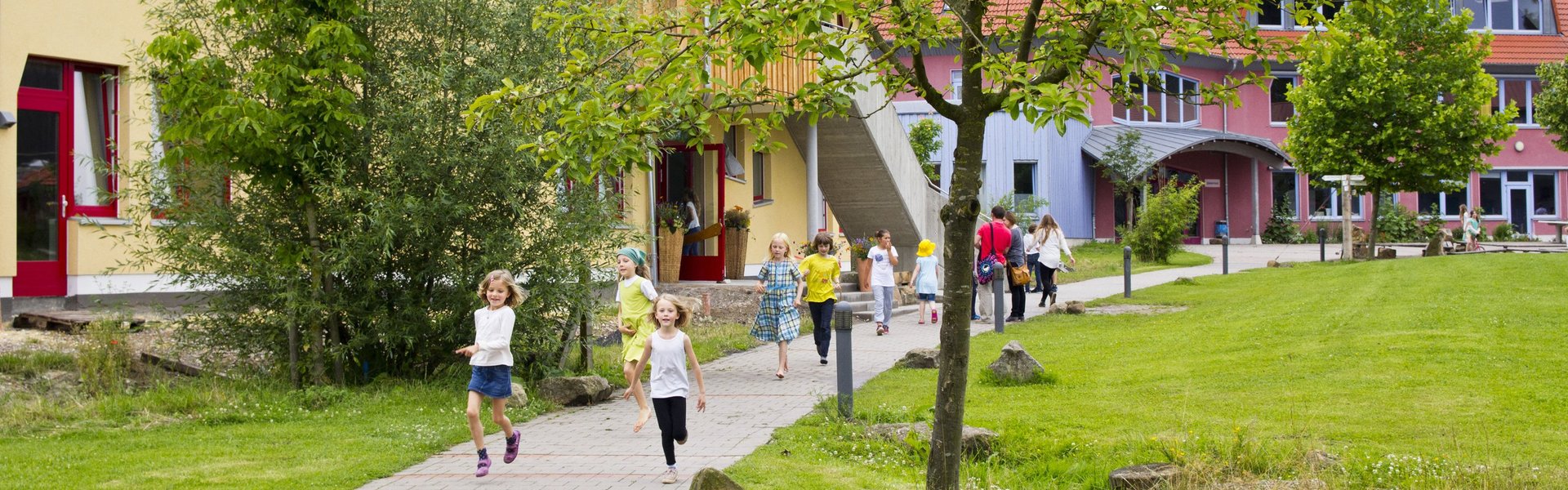An einem sonnigen Tag laufen und rennen Kinder auf einem gepflasterten Weg vor einem bunten Schulgebäude, umgeben von grünem Gras, Bäumen und Sträuchern.