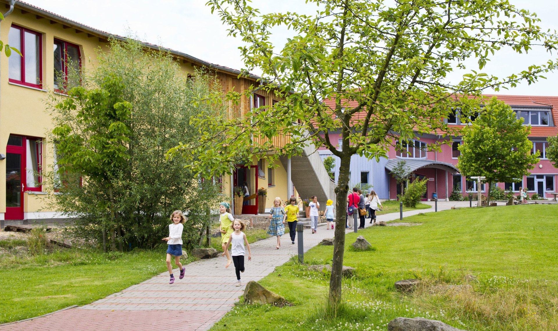 An einem sonnigen Tag laufen und rennen Kinder auf einem gepflasterten Weg vor einem bunten Schulgebäude, umgeben von grünem Gras, Bäumen und Sträuchern.
