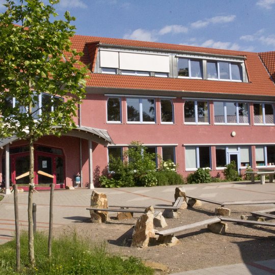 Hinter einem Spielplatz mit Holzbänken und Steinen steht ein rotes, zweistöckiges Schulgebäude mit vielen Fenstern. Dort gibt es Bäume, gepflasterte Wege und über den Köpfen einen blauen Himmel mit Wolken.