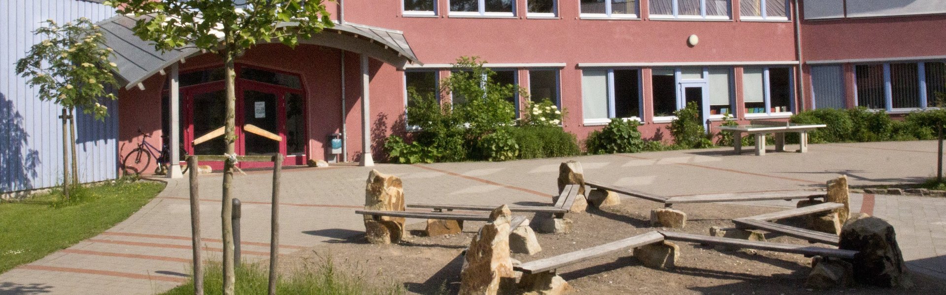 Hinter einem Spielplatz mit Holzbänken und Steinen steht ein rotes, zweistöckiges Schulgebäude mit vielen Fenstern. Dort gibt es Bäume, gepflasterte Wege und über den Köpfen einen blauen Himmel mit Wolken.