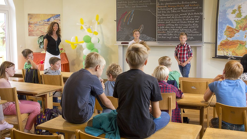 Ein Klassenzimmer, in dem Kinder an ihren Schreibtischen sitzen, zwei Jungen an der Tafel stehen, ein Lehrer in der Nähe steht und Landkarten und Luftballons den Raum schmücken.
