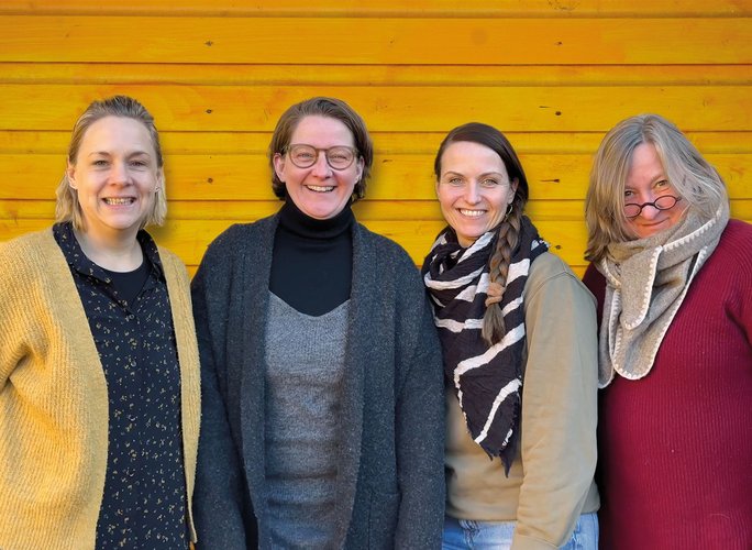 Vier lächelnde Frauen stehen nebeneinander vor einer leuchtend gelben Holzwand, tragen Pullover und Schals und haben ihre Arme umeinander gelegt.