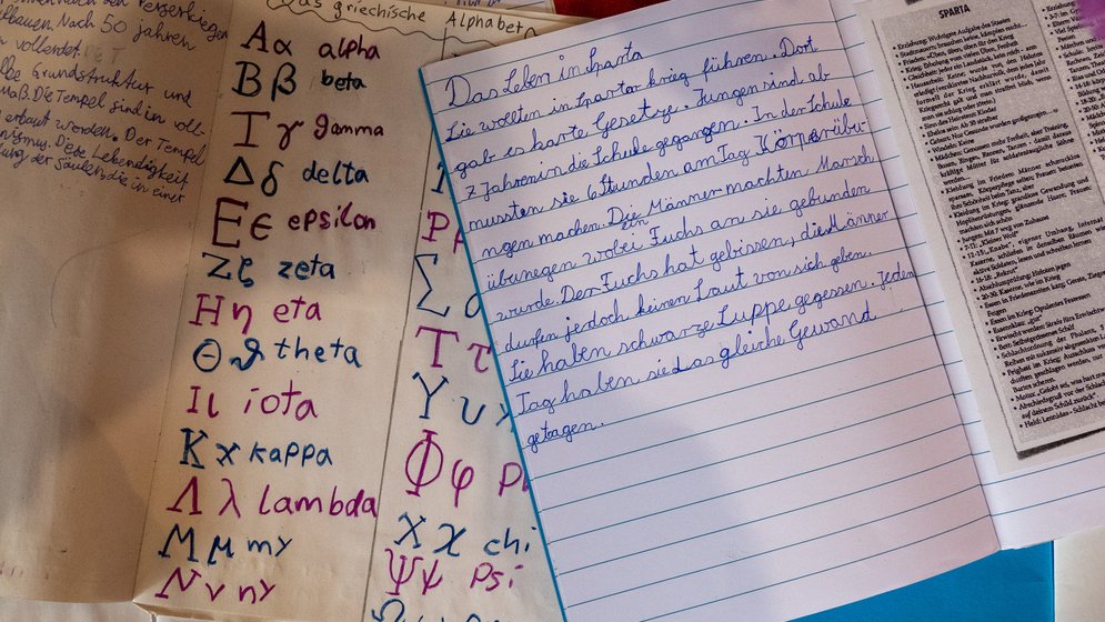 Auf einem Tisch liegen handbeschriftete Seiten mit dem griechischen Alphabet, Namen und deren Aussprache, Notizen in deutscher Schreibschrift sowie ein Zeitungsartikel.