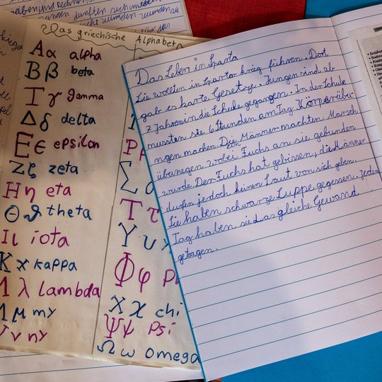 Auf einem Tisch liegen handbeschriftete Seiten mit dem griechischen Alphabet, Namen und deren Aussprache, Notizen in deutscher Schreibschrift sowie ein Zeitungsartikel.
