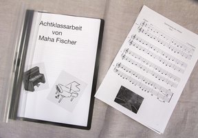 Auf einer beigen Stoffunterlage liegt neben einem gedruckten Notenblatt eine Broschüre mit dem Titel „Achtklassarbeit von Maha Fischer“ mit Klavierbegleitung.