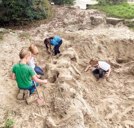 Vier Kinder graben und spielen draußen auf einer großen Sandfläche und benutzen dabei kleine Werkzeuge und ihre Hände. Bäume und Gras umgeben die Szene, und die Kinder scheinen ganz darauf konzentriert zu sein, im Sand etwas zu bauen und ihn zu erkunden.