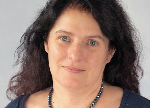 Eine Frau mit langem, welligem, dunklem Haar und blauen Augen, die ein dunkelblaues Oberteil und eine blaue Perlenkette trägt und vor einem hellen Hintergrund sanft in die Kamera lächelt.