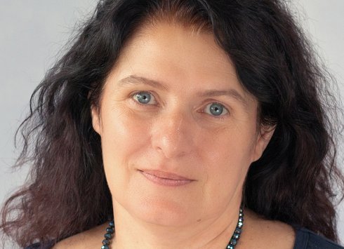Eine Frau mit langem, welligem, dunklem Haar und blauen Augen, die ein dunkelblaues Oberteil und eine blaue Perlenkette trägt und vor einem hellen Hintergrund sanft in die Kamera lächelt.