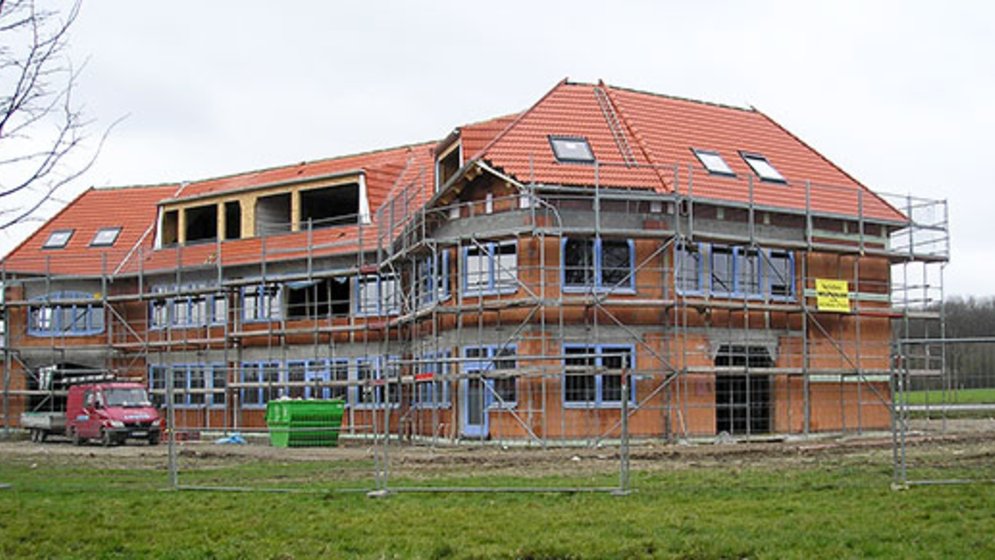 Ein zweistöckiges Backsteingebäude im Bau, mit Gerüsten, roten Dachziegeln und mehreren Fenstern. Vor dem Gebäude stehen ein roter Transporter und ein grüner Müllcontainer, und rundherum ist Gras. Der Himmel ist bewölkt.