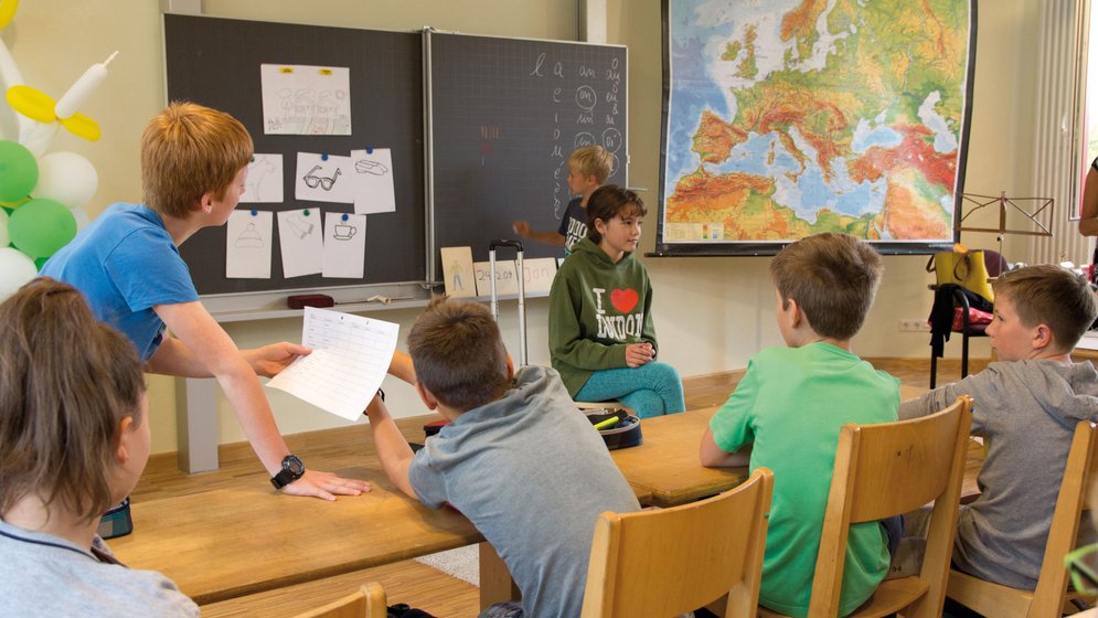 Eine Gruppe von Kindern sitzt in einem Klassenzimmer an Holzpulttischen. Ein Kind steht vorne und hält ein Blatt Papier in der Hand, während ein anderes an die Tafel schreibt. An der Wand hängen eine große Karte von Europa und einige Zeichnungen.
