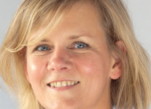 Eine Frau mit kurzen blonden Haaren und blauen Augen lächelt vor einem hellen, neutralen Hintergrund in die Kamera.