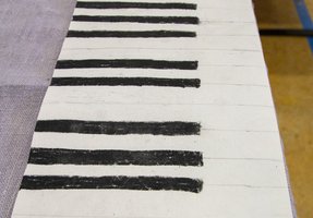 Ein Blatt Papier, auf dem waagerechte schwarze Balken in gleichmäßigen Abständen eingezeichnet sind, auf dem schwache Bleistiftlinien zu erkennen sind und dessen linke Seite teilweise von einem Stoff bedeckt ist.