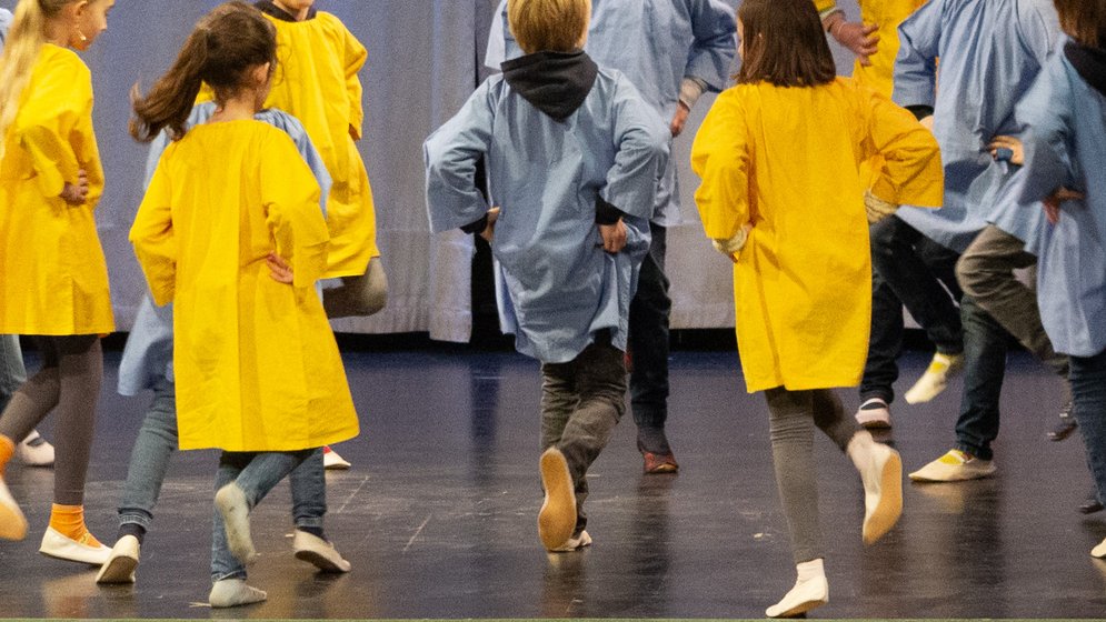 Kinder spielen draußen, lächeln und halten sich an den Händen, während sie im Kreis stehen; im Hintergrund sind Bäume und Gebäude zu sehen – an einem sonnigen Tag.