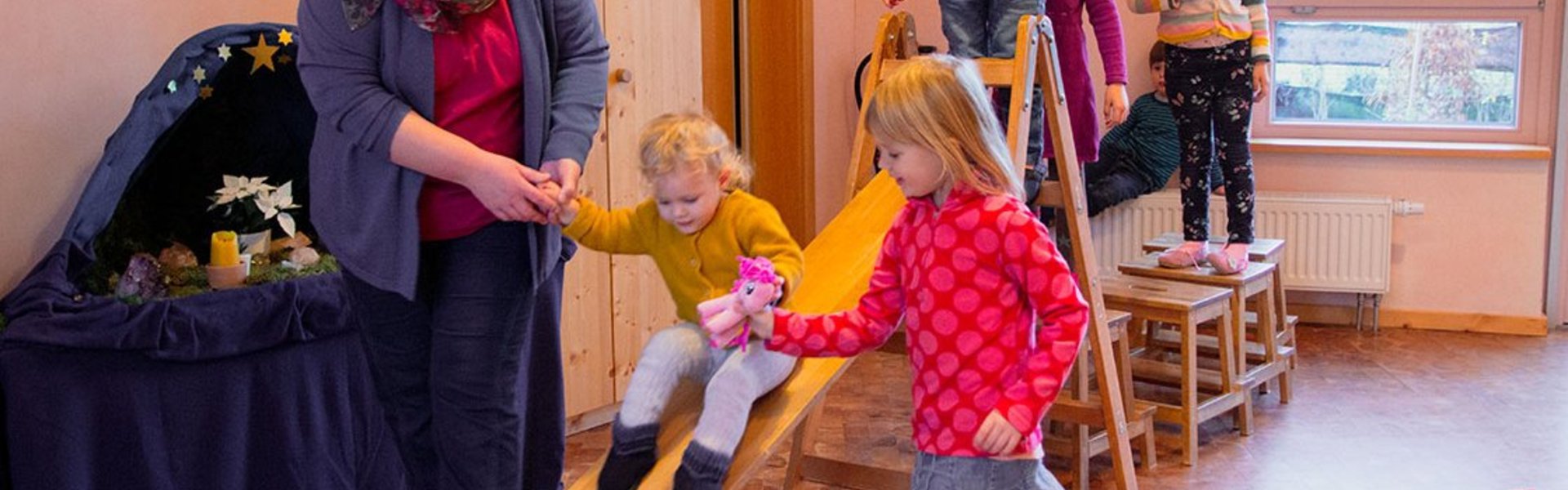 Eine Frau hilft einem kleinen Mädchen, auf einer Holzrutsche in einem Innenraum hinunterzurutschen, während andere Kinder in der Schlange warten und zuschauen. Im Hintergrund ist eine Krippe zu sehen, und durch ein Fenster sind draußen Häuser zu erkennen.