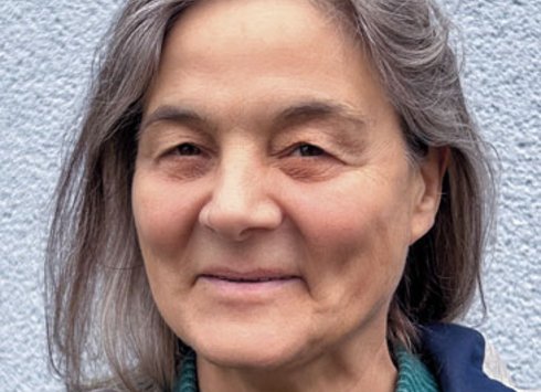Eine Frau mit glattem braunem Haar, die eine Brille mit schwarzem Rahmen und eine schwarze Halskette trägt, lächelt vor einem hellen, schlichten Hintergrund in die Kamera.