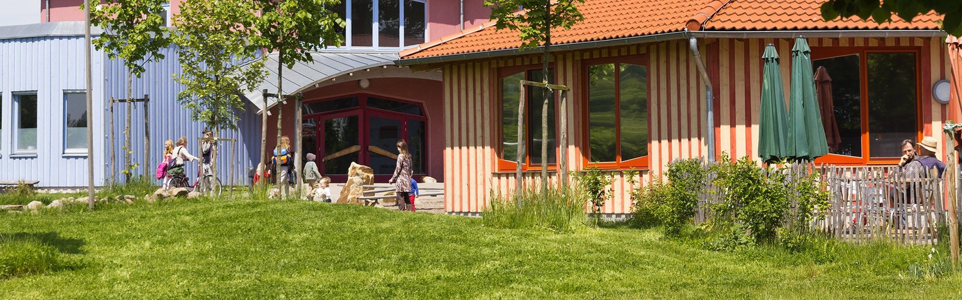 Ein farbenfrohes Gebäude mit roten Dächern, umgeben von grünem Gras und Bäumen. An einem sonnigen Tag spielen Kinder draußen in der Nähe des Eingangs.
