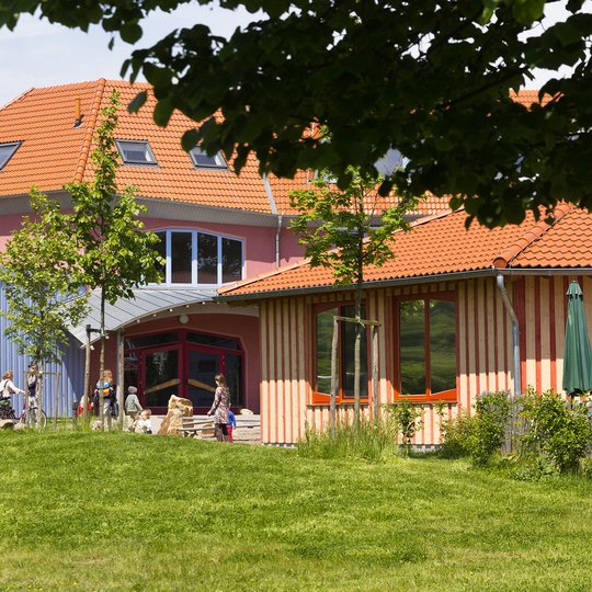 Ein farbenfrohes Gebäude mit roten Dächern, umgeben von grünem Gras und Bäumen. An einem sonnigen Tag spielen Kinder draußen in der Nähe des Eingangs.