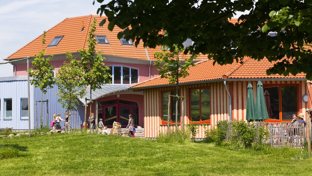 Ein farbenfrohes Gebäude mit roten Dächern, umgeben von grünem Gras und Bäumen. An einem sonnigen Tag spielen Kinder draußen in der Nähe des Eingangs.