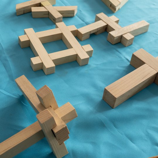 Auf einer hellblauen Stofffläche sind mehrere Holzklötze angeordnet. Die Klötze sind so ineinander gesteckt, dass sie verschiedene geometrische Formen und Muster bilden.