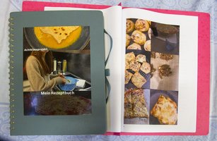Neben einem aufgeschlagenen Kochbuch, in dem verschiedene Backwaren auf einem rosa-weiß gemusterten Hintergrund abgebildet sind, liegt ein grünes Spiralheft mit der Aufschrift „Achtklassprojekt Mein Rezeptbuch“.