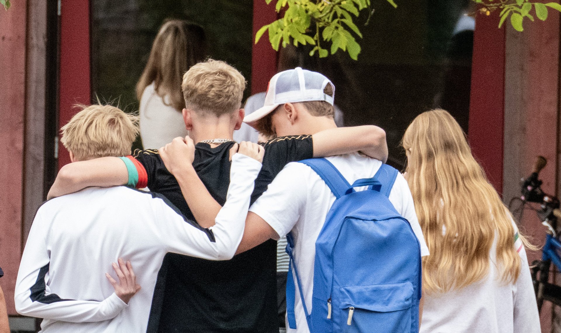 Vier Jugendliche, von hinten gesehen, gehen dicht beieinander und haben die Arme um die Schultern der anderen gelegt. Einer trägt einen blauen Rucksack, ein anderer hat langes blondes Haar. Im Hintergrund sind Bäume und ein rotes Gebäude zu sehen.