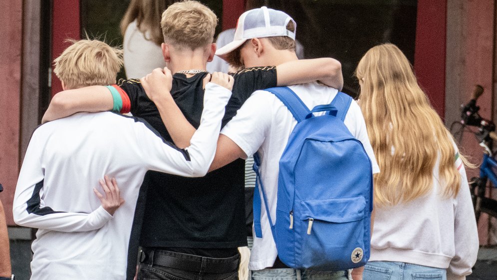 Vier Jugendliche, von hinten gesehen, gehen dicht beieinander und haben die Arme um die Schultern der anderen gelegt. Einer trägt einen blauen Rucksack, ein anderer hat langes blondes Haar. Im Hintergrund sind Bäume und ein rotes Gebäude zu sehen.
