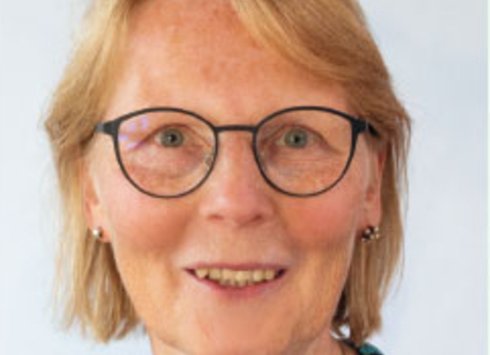 Eine Frau mittleren Alters mit kurzen blonden Haaren, einer runden Brille, kleinen Ohrringen und einem bunt gemusterten Oberteil lächelt vor einem schlichten, hellen Hintergrund in die Kamera.