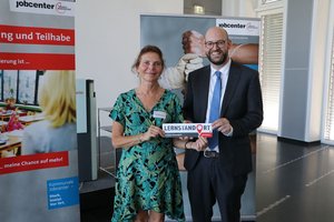 Zwei Personen stehen in einem Raum und halten ein Schild mit der Aufschrift „Lernstandort“ in den Händen. Die Frau trägt ein grün gemustertes Kleid, der Mann einen Anzug und eine Krawatte. Hinter ihnen sind Banner des Jobcenters und ein Foto von Händen zu sehen, die Teig kneten.