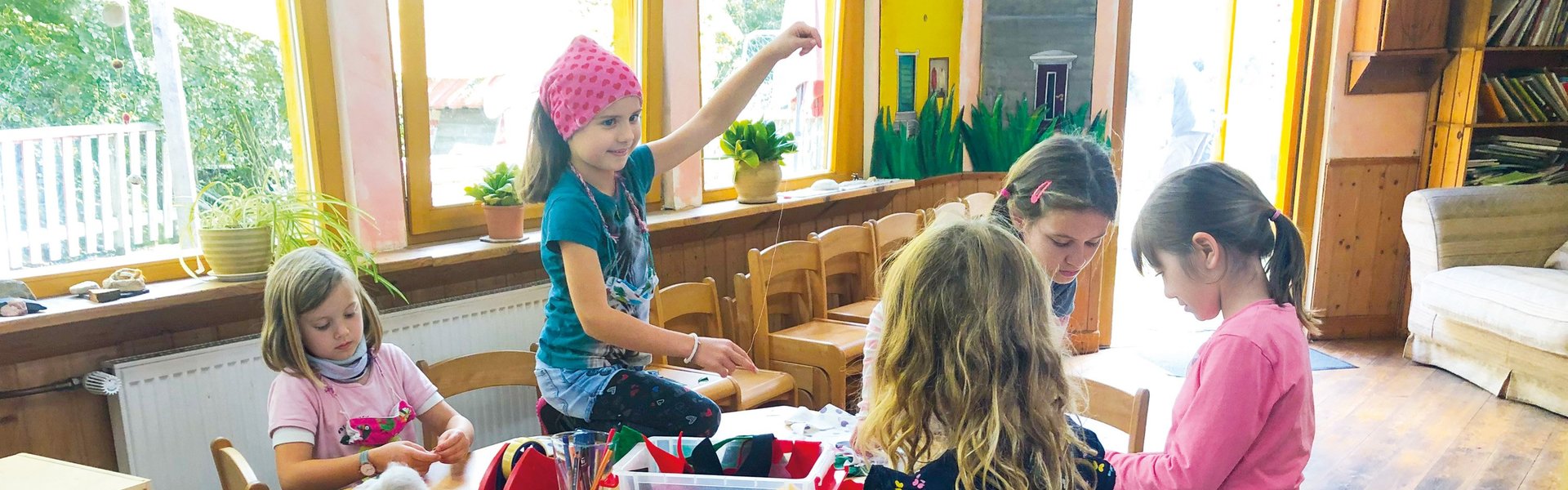 Fünf kleine Kinder sitzen in einem hell erleuchteten Klassenzimmer um einen Tisch herum und basteln mit bunten Materialien. Im Hintergrund sind Regale mit Büchern und große Fenster zu sehen.