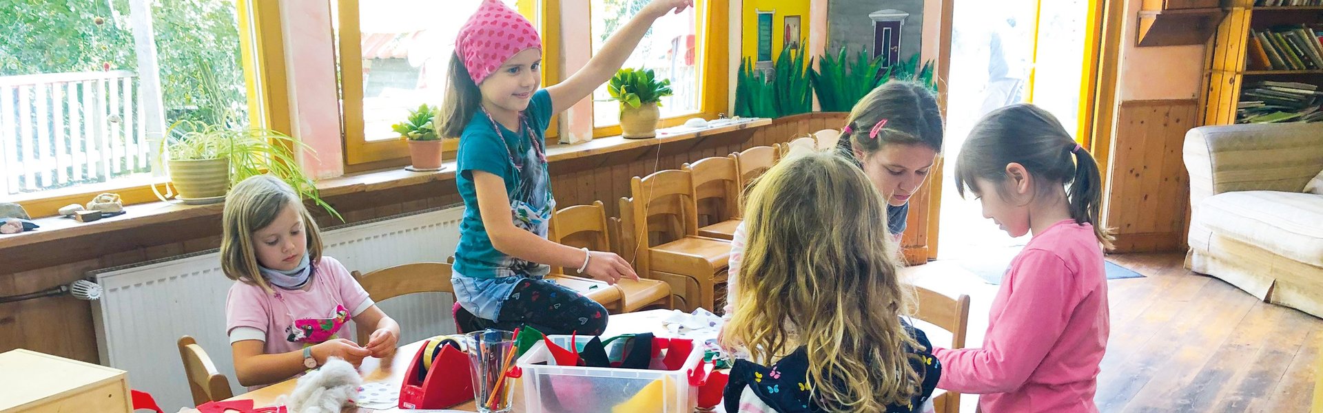 Fünf kleine Kinder sitzen in einem hell erleuchteten Klassenzimmer um einen Tisch herum und basteln mit bunten Materialien. Im Hintergrund sind Regale mit Büchern und große Fenster zu sehen.