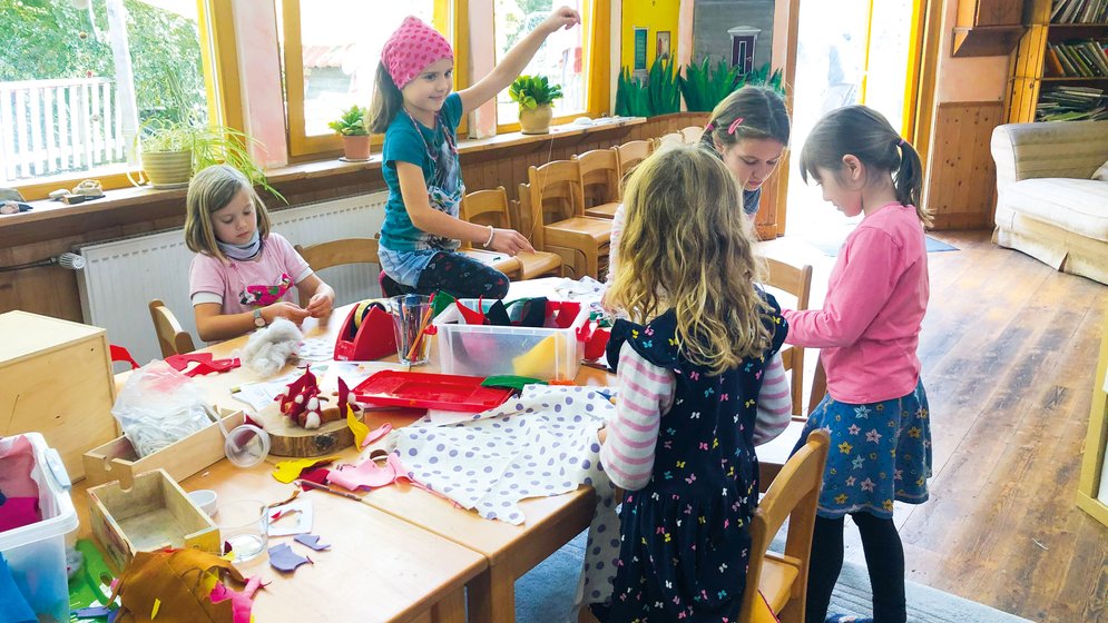 Fünf kleine Kinder sitzen in einem hell erleuchteten Klassenzimmer um einen Tisch herum und basteln mit bunten Materialien. Im Hintergrund sind Regale mit Büchern und große Fenster zu sehen.
