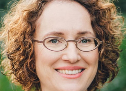 Eine Frau mit lockigem, hellbraunem Haar, die eine Brille und ein weißes Hemd trägt, lächelt im Freien vor einem grünen, verschwommenen Hintergrund.