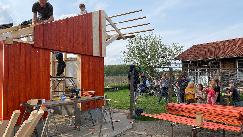 Ein noch nicht ganz fertiggestellter Holzschuppen mit roter Verkleidung, freiliegenden Balken und überall verstreutem Werkzeug. Frisch gestrichene Bretter trocknen auf Sägeböcken. Im Hintergrund sind ein Maschendrahtzaun und ein Haus mit rotem Dach zu sehen.