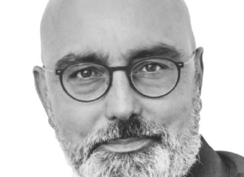 Ein kahlköpfiger Mann mit Brille und grauem Bart, der ein Sakko und ein weißes Hemd trägt und vor einem schlichten weißen Hintergrund mit ausdruckslosem Gesichtsausdruck direkt in die Kamera blickt.