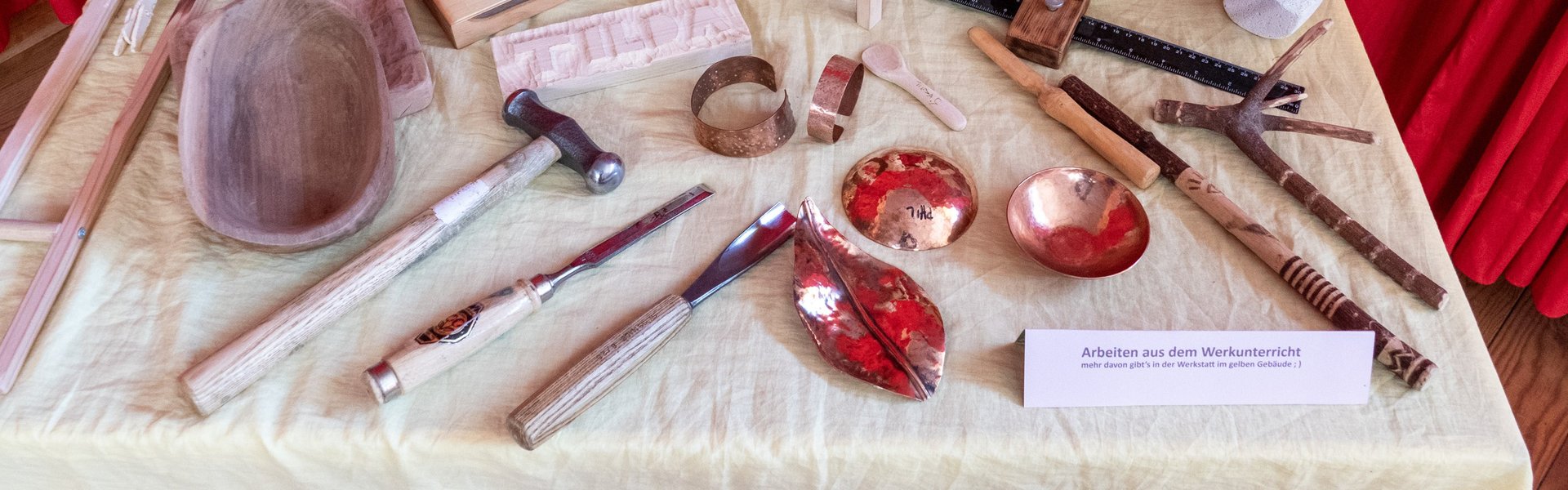 Ein Ausstellungs-Tisch mit Holzskulpturen, Werkzeugen, geschnitzten Schalen und Metallgegenständen vor einem roten Vorhang, auf dem verschiedene handgefertigte Gegenstände präsentiert werden.