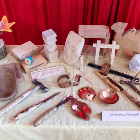 Ein Ausstellungs-Tisch mit Holzskulpturen, Werkzeugen, geschnitzten Schalen und Metallgegenständen vor einem roten Vorhang, auf dem verschiedene handgefertigte Gegenstände präsentiert werden.