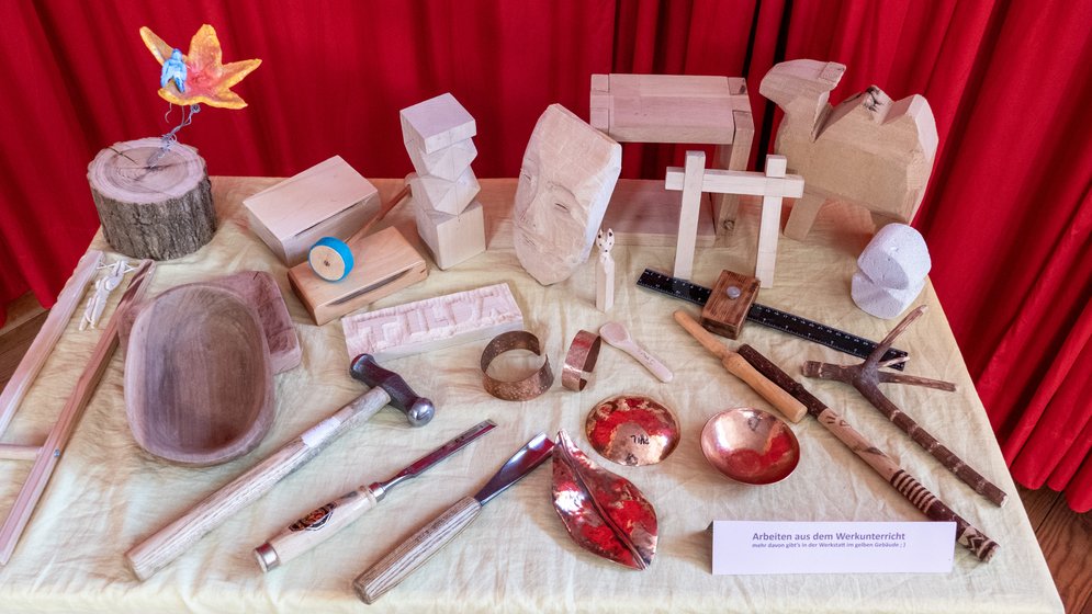 Ein Ausstellungs-Tisch mit Holzskulpturen, Werkzeugen, geschnitzten Schalen und Metallgegenständen vor einem roten Vorhang, auf dem verschiedene handgefertigte Gegenstände präsentiert werden.